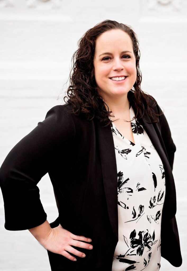 A headshot of Lindsey Baker. She is a white woman with brown, curly hair past her shoulders. She wears a white button-down shirt with black flowers on it and a black blazer. Her hands are on her hips. The background is white.