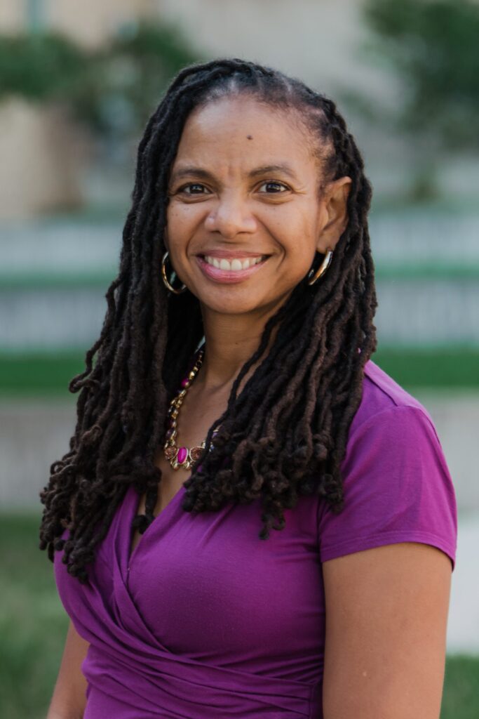 Dr. Kimberly Moffitt, a middle-aged-looking Black woman with her hair down in locs. She wears gold hoop earrings and a darker magenta short-sleeved shirt. The background looks like artistically blurry trees in front of a building.