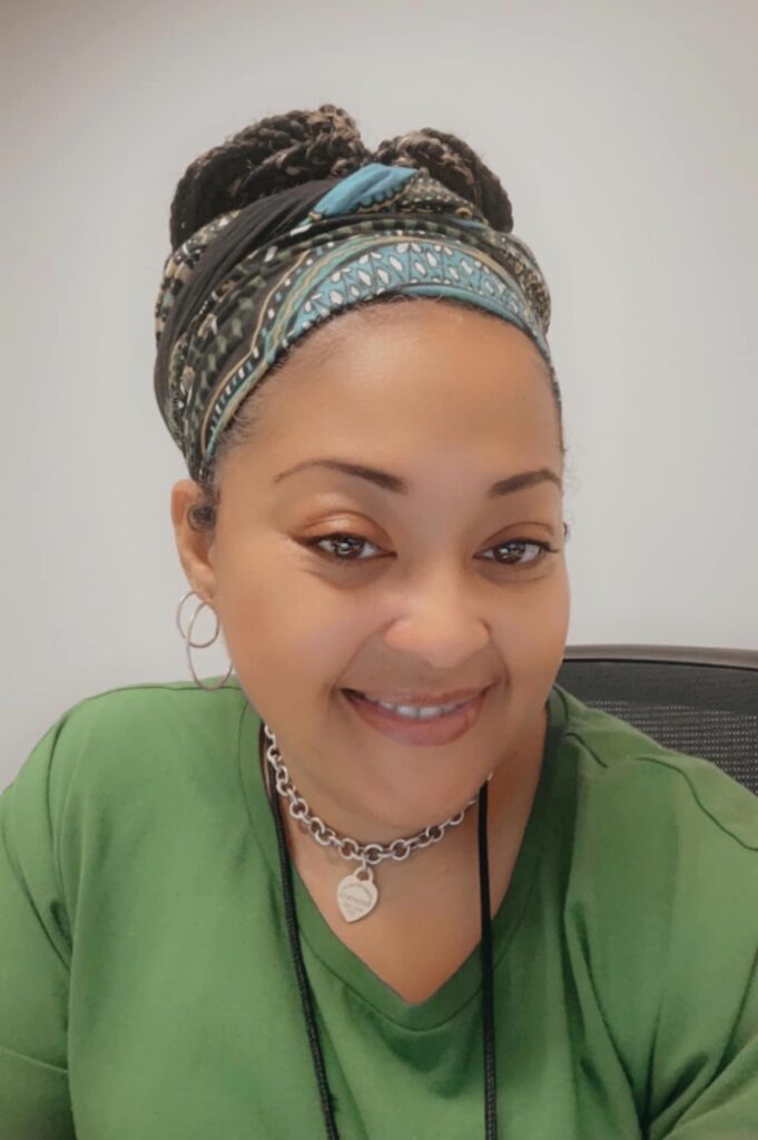 Mariam Spalding is a Black woman who wears a long sleeved green shirt. She wears her hair up in locs with a black and turquoise patterned head scarf. She has a silver choker necklace with a heart.