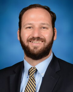 A professional photograph of Sam Manelski, a middle-aged white man with brown hair and a beard and mustache. He sits or stands in front of a blue backdrop. He wears a black or navy blue blazer, yellow and black or navy blue striped tie, and a light blue button down shirt.
