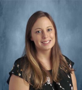 A professional photograph of Lindsay Matthews, a young or middle-aged white woman with straight and dirty blonde hair that she wears down. She sits or stands in front of a blue backdrop. She wears a black or navy blue top or dress with a floral pattern on it.